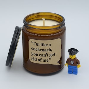 May include: A brown glass candle jar with a white candle inside. The jar has a label that reads "I'm like a cockroach, you can't get rid of me." A small Lego figure with a pirate hat and a fox logo on its shirt is standing next to the candle.