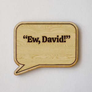 May include: Wooden speech bubble-shaped sign with the phrase "Ew, David!" in dark brown lettering. The sign has a light wood grain pattern and a dark brown border, perfect for home decor or gifting.