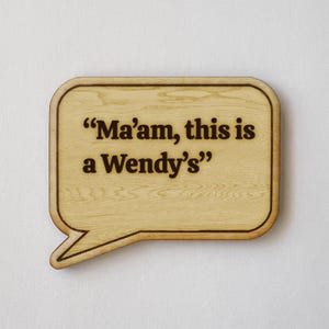 May include: Wooden speech bubble-shaped sign with the phrase "Ma'am, this is a Wendy's" in dark brown lettering. The sign has a light wood grain finish and a dark brown border, perfect for home decor.
