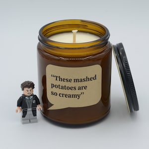 May include: A brown glass candle jar with a beige label that reads "These mashed potatoes are so creamy." The candle is lit and the jar has a black lid.