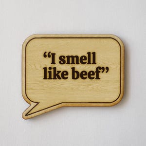 May include: A wooden speech bubble-shaped sign with the phrase "I smell like beef" in dark brown lettering. The sign has a light wood grain finish and a darker brown border, suitable for home decor.