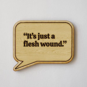 May include: Wooden speech bubble-shaped sign with the quote "It's just a flesh wound" in dark brown lettering. The sign has a light wood grain finish and a dark brown border, perfect for home decor.