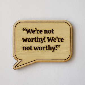 May include: A wooden speech bubble-shaped sign with the phrase "We're not worthy! We're not worthy!" in dark brown lettering. The sign has a light wood grain finish and a dark brown border.