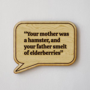 May include: Wooden speech bubble-shaped sign with the quote "Your mother was a hamster, and your father smelt of elderberries" engraved in dark brown. The sign has a light brown wood grain finish and a dark brown border.