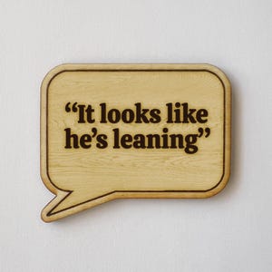 May include: Wooden speech bubble-shaped sign with the quote "It looks like he's leaning" in dark brown lettering. The sign has a light wood grain pattern and a rounded border.