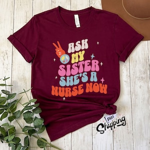 May include: Burgundy t-shirt with a peace sign and the text "Ask my sister she's a nurse now" in a retro font.