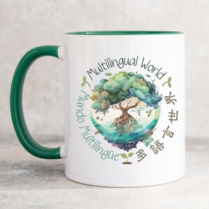 May include: White ceramic mug with a green rim and handle. The mug features a colourful illustration of a tree with roots growing into a globe. The text "Multilingual World" is written around the tree in a circle, along with the words "Mundo Multilingüe" and "多言語世界" in different languages.