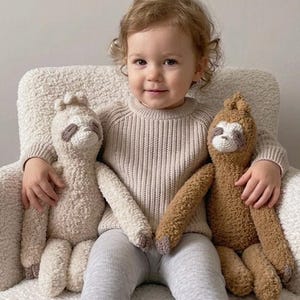 May include: A child sits in a white, fluffy armchair, holding two crocheted sloth plush toys. The sloths are beige and brown, with detailed facial features. The child wears a beige sweater and gray leggings.