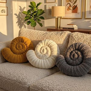 Pode incluir: Três almofadas em forma de espiral nas cores amarelo mostarda, creme e cinza escuro, cada uma com uma superfície texturizada e felpuda. Estas almofadas estão dispostas num sofá texturizado de cor creme, exibindo um design de concha.