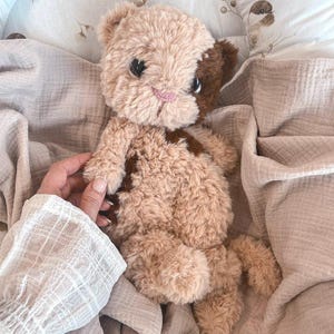 May include: A plush stuffed animal resembling a cat with soft, textured beige and dark brown fur. The toy features large, dark, glossy eyes and a small, embroidered pink nose. It has a rounded head, small ears, and a soft, fuzzy body.