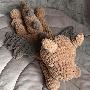 May include: A plush dragon toy crafted from tan and dark gray yarn. The creature features a textured, knotted surface, prominent dark gray wings, and a series of small dark gray spikes running down its back. It has two small ears and a rounded head.