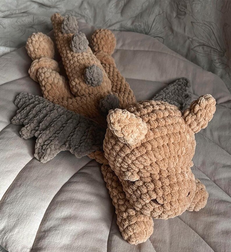 May include: A plush dragon toy crafted from tan and dark gray yarn. The creature features a textured, knotted surface, prominent dark gray wings, and a series of small dark gray spikes running down its back. It has two small ears and a rounded head.