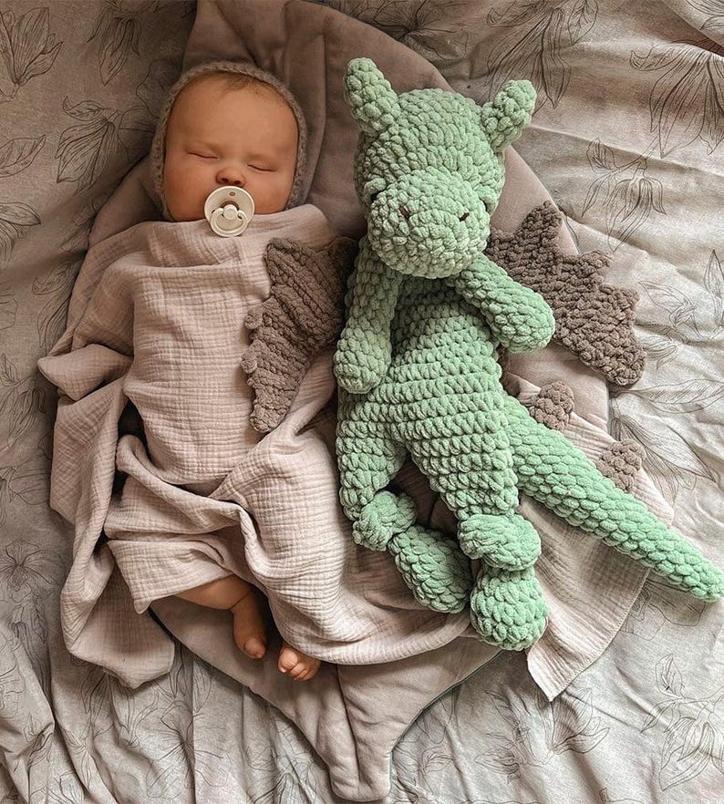 May include: A sleeping infant wrapped in a light beige muslin swaddle with a matching bonnet and a white pacifier, positioned next to a light green crocheted dragon plush toy featuring textured yarn and dark brown felt-like wing accents.