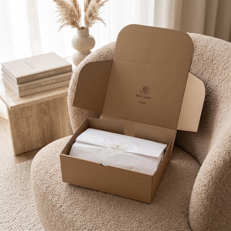 May include: A brown cardboard shipping box with an open lid, featuring the BalticTinyKnit logo printed on the inside. Inside the box, white tissue paper is neatly folded and secured with a small rectangular sticker bearing the same brand logo.
