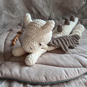May include: A crocheted dragon plush toy made of thick, soft white yarn with dark gray wings and a row of dark gray spikes along its back. The creature has two pointed ears and is positioned in a resting pose. A small wooden bead pacifier clip is nearby.