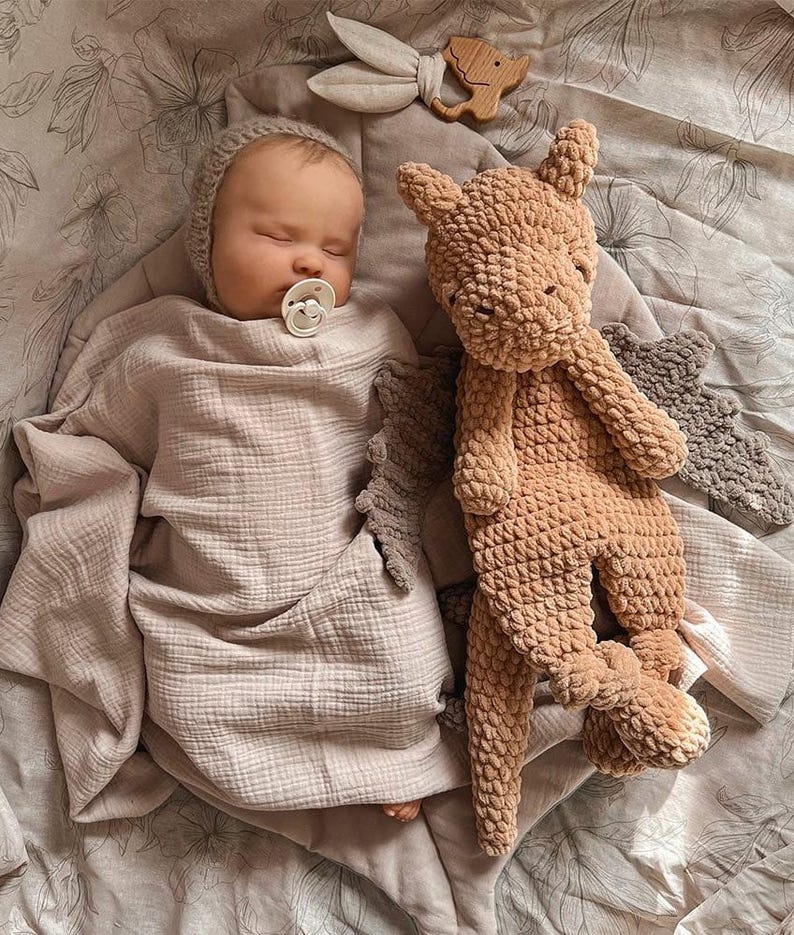 May include: A sleeping infant wearing a light beige knit bonnet and a white pacifier, wrapped in a soft, light beige muslin swaddle. Beside the infant is a plush, textured tan dragon toy with dark gray wings and a small wooden animal-shaped teether.