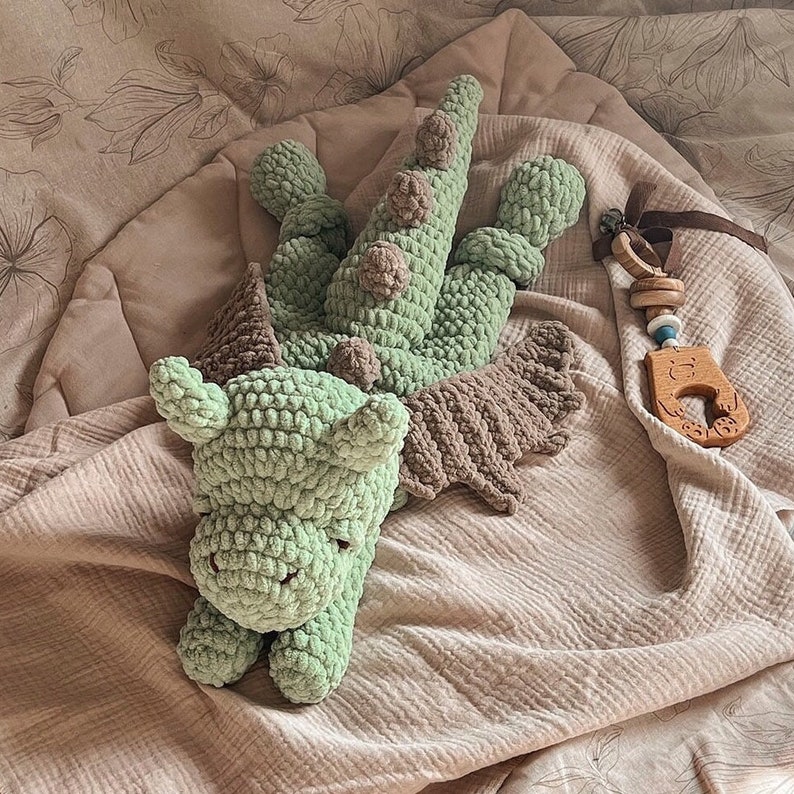 May include: A light green crocheted dragon plush toy with brown wings and brown spikes along its back. Beside the dragon lies a wooden teether toy featuring a carved bear face, a brown ribbon, and small wooden rings attached to a blue and white bead.