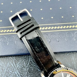May include: A black rubber watch strap with a silver-toned metal buckle. The strap features a smooth, glossy finish on the main band and a matte texture on the keeper loops. The watch case is partially visible at the bottom with a black dial.