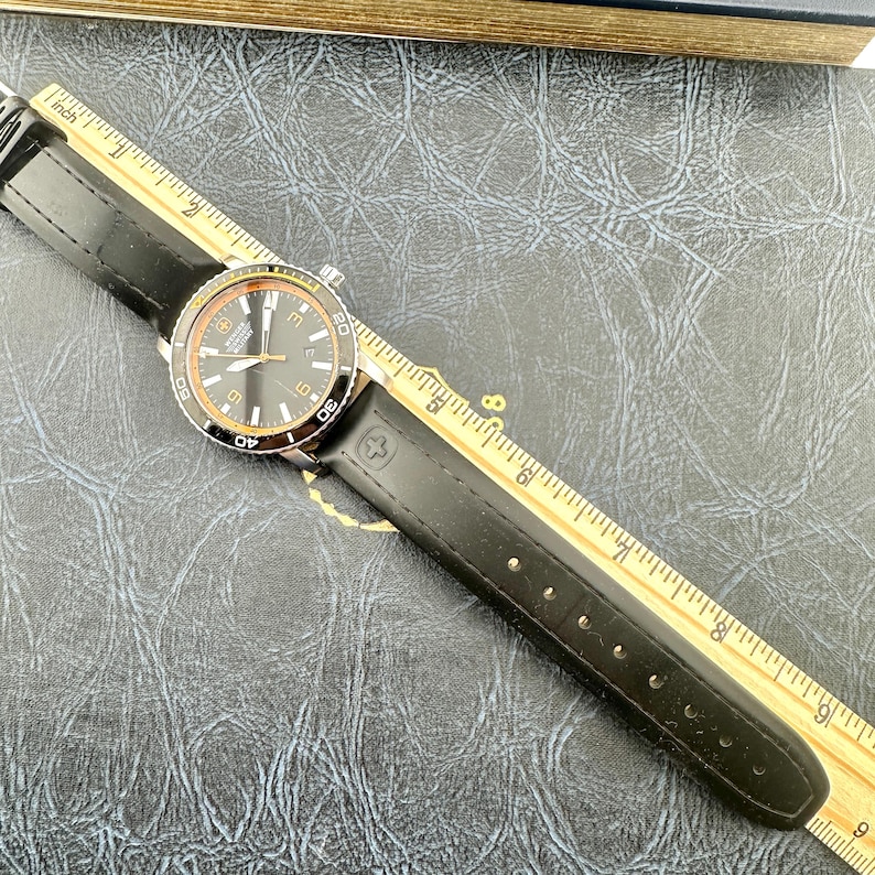 May include: A Wenger Swiss Military wristwatch with a black dial, silver-tone hands, and a black leather strap. The watch is positioned next to a yellow wooden ruler, showing a total length of approximately 9.5 inches from end to end.