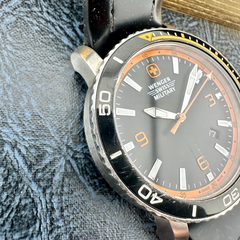 May include: A Wenger Swiss Military wristwatch with a black dial, orange numerals at 3, 6, and 9 o'clock, and a date window at 3 o'clock. The watch features a black rotating bezel with white markings, a black silicone strap, and silver-tone hands.