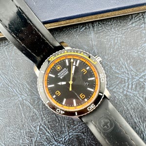 May include: A Wenger Swiss Military wristwatch featuring a black dial with orange accents, luminous hands, and a date window at the three o'clock position. The watch has a black rotating bezel with white numerals and a black silicone strap with a logo.