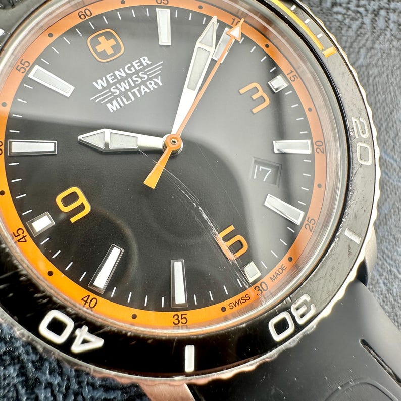 May include: A Wenger Swiss Military wristwatch with a black dial, orange inner bezel, and luminous hands. The face features the Wenger logo, Arabic numerals at 3, 6, and 9, a date window at 3 o'clock, and a black rotating outer bezel.
