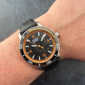 May include: A Wenger Swiss Military wristwatch featuring a black dial with orange accents, luminous hands, and large orange numerals at the three, six, and nine o'clock positions. The watch has a black rotating bezel and a black silicone strap.