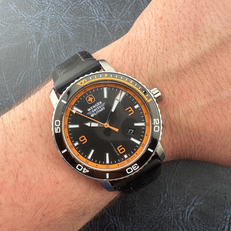 May include: A Wenger Swiss Military wristwatch featuring a black dial with orange accents, luminous hands, and large orange numerals at the three, six, and nine o'clock positions. The watch has a black rotating bezel and a black silicone strap.
