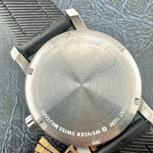 May include: A stainless steel watch case back with a circular brushed finish, featuring the text WENGER SWISS MILITARY, SAPPHIRE CRYSTAL, and WATER RESISTANT 100M. The watch is attached to a black textured rubber strap with a grooved pattern.