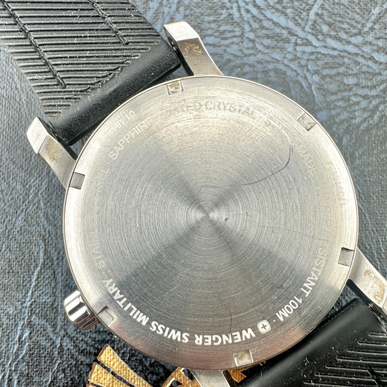 May include: A stainless steel watch case back with a circular brushed finish, featuring the text WENGER SWISS MILITARY, SAPPHIRE CRYSTAL, and WATER RESISTANT 100M. The watch is attached to a black textured rubber strap with a grooved pattern.