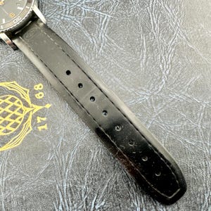May include: A black watch strap with a smooth finish and a series of circular adjustment holes, attached to a watch case with a dark dial featuring white markers and the numbers 17 and 68 printed in gold on the surface beneath the strap.