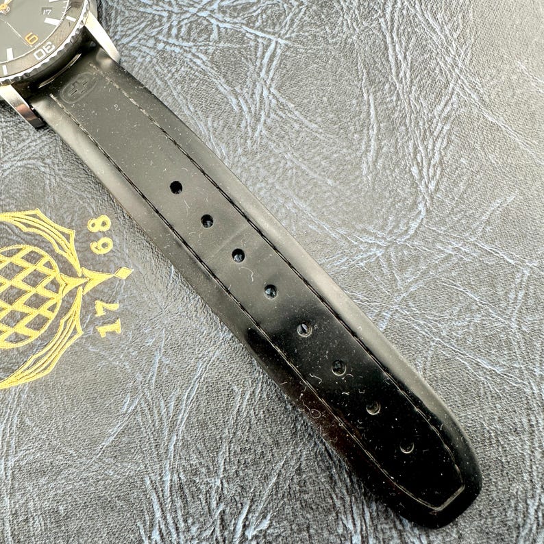 May include: A black watch strap with a smooth finish and a series of circular adjustment holes, attached to a watch case with a dark dial featuring white markers and the numbers 17 and 68 printed in gold on the surface beneath the strap.
