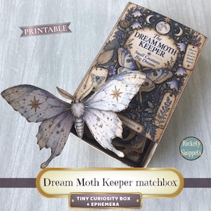 Peut inclure: Une boîte d'allumettes rectangulaire étiquetée The Dream Moth Keeper avec le texte Small Creatures Big Dreams. Un grand papillon en papier détaillé avec des motifs d'étoiles sur ses ailes repose dessus. La boîte présente des illustrations célestes.