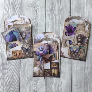 Puede incluir: Tres organizadores de bolsillo de papel decorativos con un tema de hadas vintage. Cada bolsillo presenta ilustraciones de hadas, mariposas y flores en tonos morados, azules y crema. Los bolsillos est&aacute;n llenos de tarjetas y etiquetas m&aacute;s peque&ntilde;as.