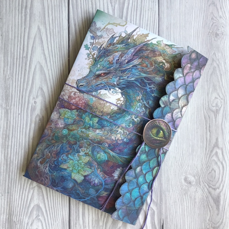 May include: A blue and purple dragon with intricate details is featured on the cover of a journal. The journal has a decorative, scalloped edge and a wax seal with a dragon eye.