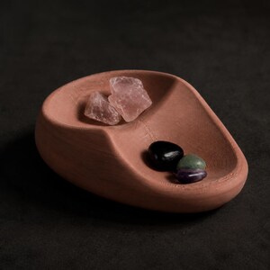 May include: A terracotta-colored, teardrop-shaped tray with a textured, ridged interior surface. The tray holds two translucent, pale pink raw crystal chunks in one section and two smooth, polished stones in black and green in the other.
