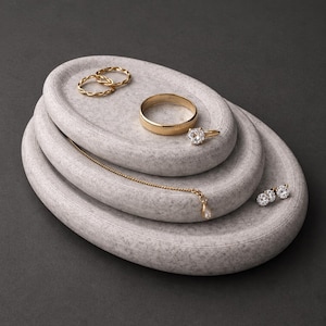 May include: Gold jewellery displayed on three oval, grey stone trays. The jewellery includes two twisted rings, a classic band, a diamond engagement ring, a delicate necklace, and a pair of diamond stud earrings. The background is a dark grey.