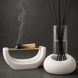 Può includere: Un porta incenso in pietra bianca con un'asta orizzontale in ottone che sostiene un bastoncino di palo santo fumante, accanto a una base per diffusore in pietra bianca con un cilindro di vetro trasparente pieno di liquido e bastoncini neri.