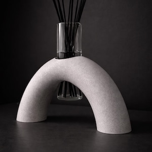 Könnte beinhalten: Ein modernes Wohnaccessoire mit einer klaren Glasvase, gefüllt mit schwarzen Schilfhalmen, auf einem hellgrauen, bogenförmigen Dekorationsobjekt. Das minimalistische Design vor dunklem Hintergrund.