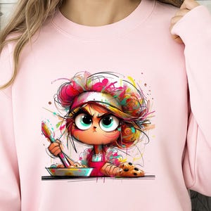 May include: Pink sweatshirt featuring a cartoon illustration of a chef with an angry expression. The chef is wearing a white hat and red apron, holding a spoon over a bowl. Colorful paint splatters and a plate of cookies complete the design.
