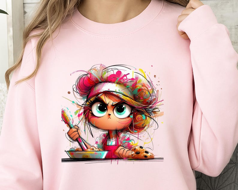 May include: Pink sweatshirt featuring a cartoon illustration of a chef with an angry expression. The chef is wearing a white hat and red apron, holding a spoon over a bowl. Colorful paint splatters and a plate of cookies complete the design.