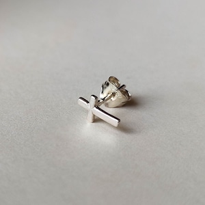 May include: A silver cross stud earring. The cross is simple and minimalist, with a slightly textured finish.