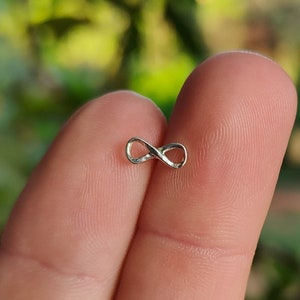 May include: A small, silver-colored infinity symbol. The figure eight shape is delicate and appears to be made of metal. The background is blurred, with green and yellow tones suggesting foliage.