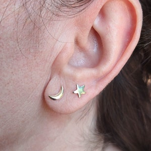 May include: A person's ear with two small gold earrings. One earring is a crescent moon shape and the other is a five-pointed star.
