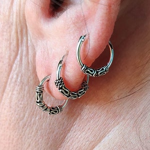 May include: Three silver hoop earrings with intricate, detailed designs. The earrings are worn in a person's ear.