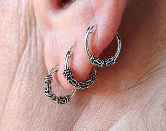 May include: Three silver hoop earrings with intricate, detailed designs. The earrings are worn in a person's ear.