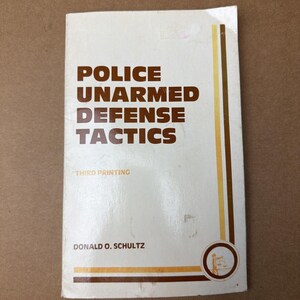 Pode incluir: Um livro de capa mole intitulado Police Unarmed Defense Tactics de Donald O. Schultz. A capa branca apresenta texto castanho, uma nota de terceira impressão e um gráfico circular com uma borda de riscas douradas e castanhas.