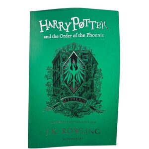 Puede incluir: Un libro de tapa dura verde titulado Harry Potter and the Order of the Phoenix, Hogwarts House Edition. La portada presenta una ilustración verde oscuro de un edificio y un emblema de fénix con la palabra Slytherin, de J.K. Rowling.