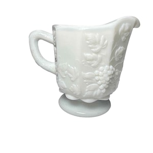 Peut inclure: Un pot à lait en verre opalin blanc présentant un motif en relief de grappes de raisin et de feuilles sur l'extérieur. Le pot possède une anse incurvée, une base festonnée et un bec verseur, le tout en verre opaque semi-translucide.