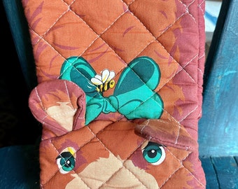 May include: A brown quilted oven mitt with a cartoon deer face. The deer has a green bow with a bee on it and a red nose.